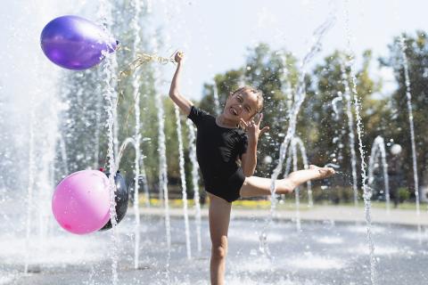 girl balancing on one foot in spray of spray park holding balloons smiling