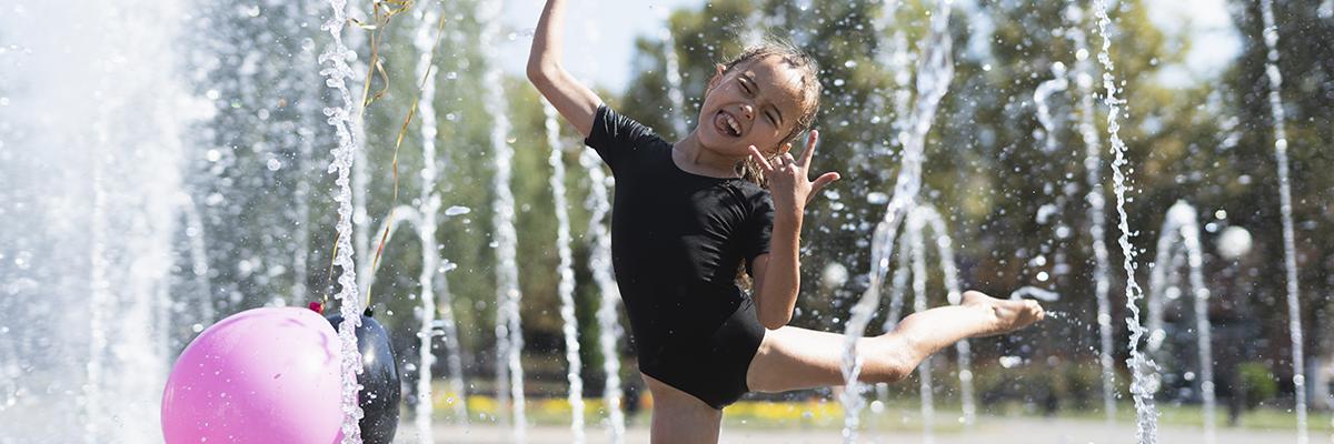 girl balancing on one foot in spray of spray park holding balloons smiling